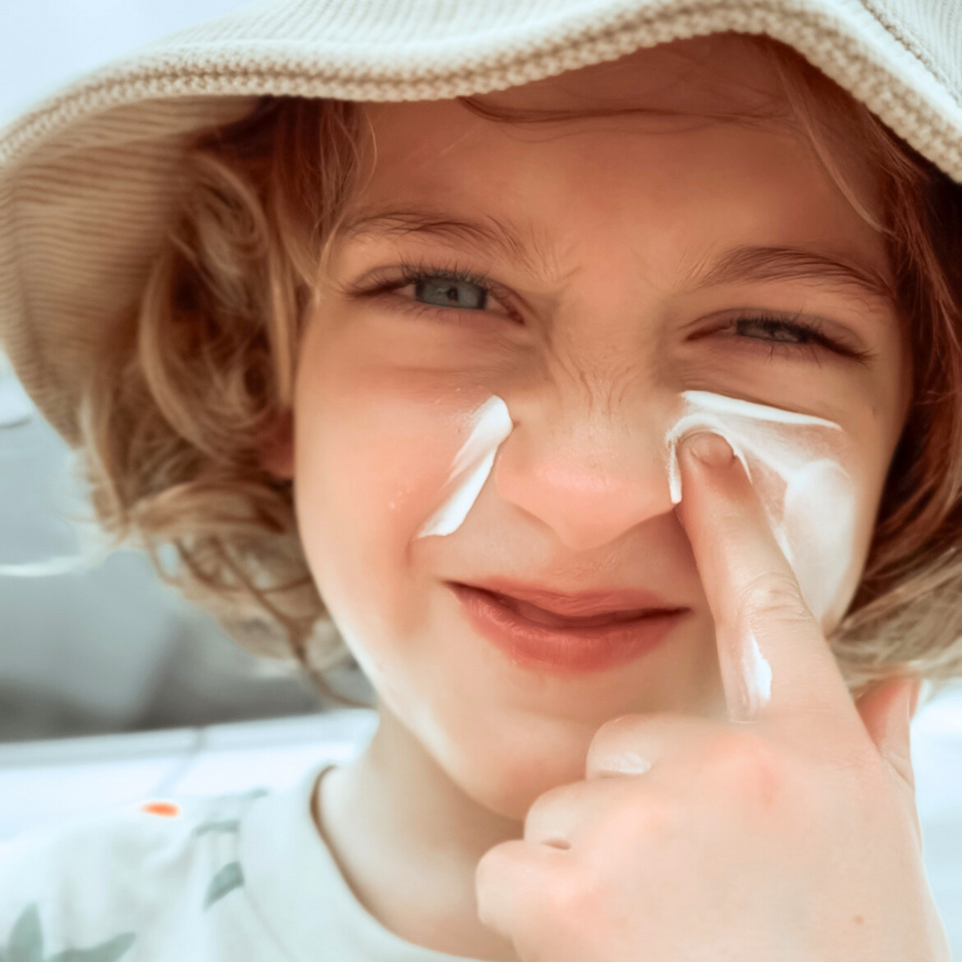 A blonde haired boy wearing a hat, screws up his nose whilst applying sunscreen to his face. He doesn't look very happy. He needs a SUNNYBOD sunscreen applicator to apply it more easily.   