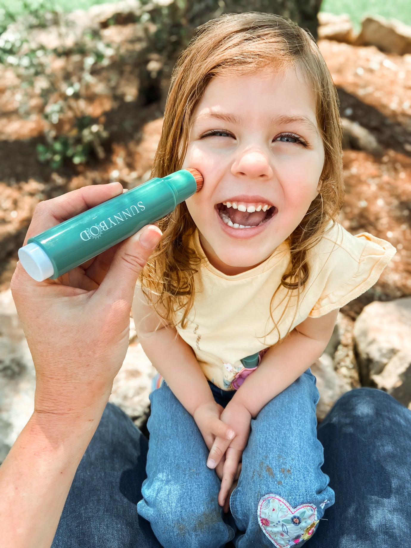 The picture is of a young girl, who appears to be very happy and is smiling cheekily. The child has wavy brown hair. There is an adult hand holding a SUNNYBOD™ Refillable Sunscreen Brush applicator and is applying sunscreen to the child's face. 