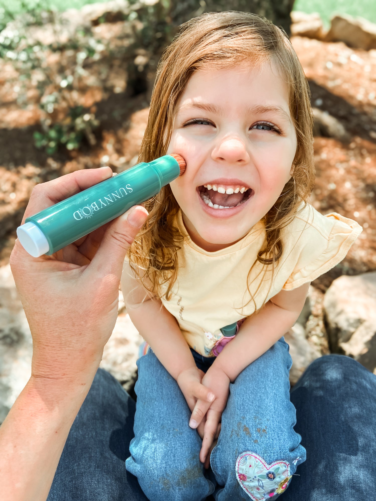 The picture is of a young girl, who appears to be very happy and is smiling cheekily. The child has wavy brown hair. There is an adult hand holding a SUNNYBOD™ Refillable Sunscreen Brush applicator and is applying sunscreen to the child's face. 