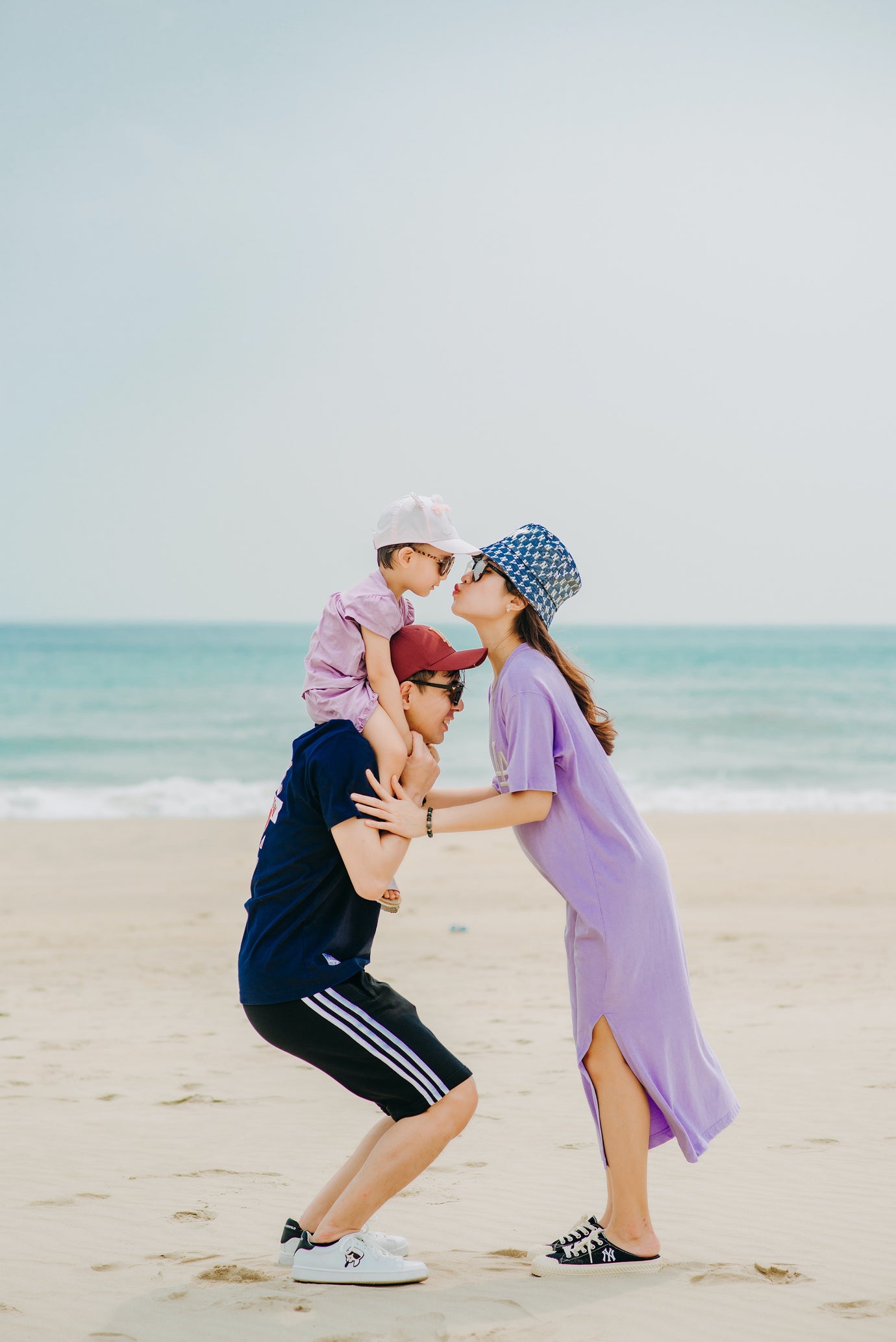 Image of a child on their dads shoulders. Their mother is standing in front of them leaning forward to kiss their child on the cheek. They are dressed in covered beach wear for sun protection. Image credit: Trần Long