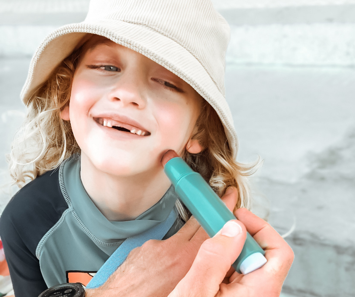 The picture is of a young boy, who appears to be very happy and is smiling widely. The child is wearing a light-coloured bucket hat and has long, curly blonde hair. There is an adult hand holding a SUNNYBOD™ Refillable Sunscreen Brush and is applying sunscreen to the child's face. The child is also wearing a dark-colored swim shirt with some design on it. They are on grey steps.