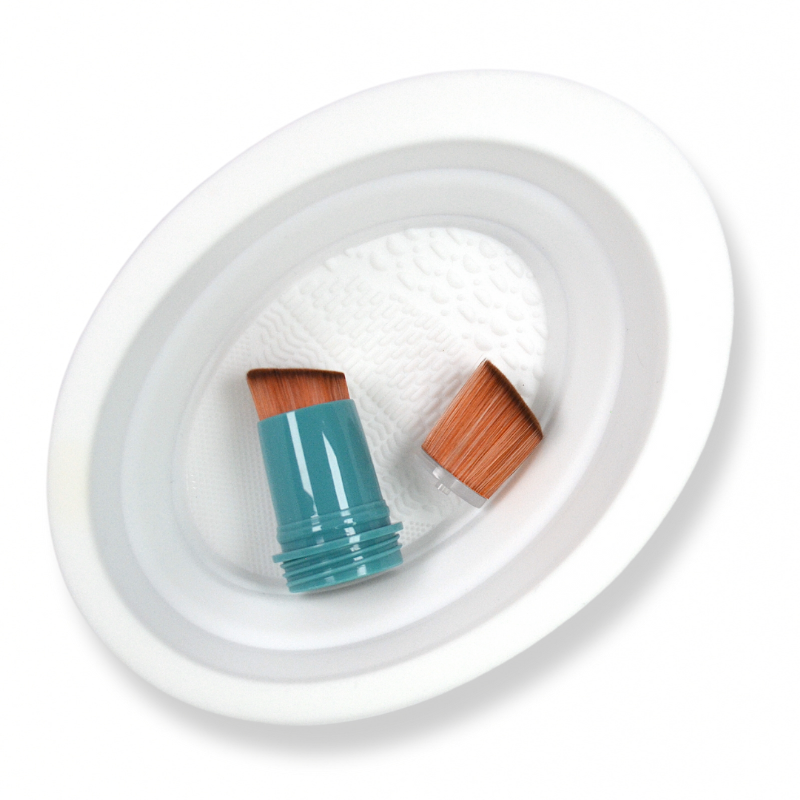 A picture of a white SUNNYBOD™ Silicone Brush Cleaning Bowl. The picture is a flat lay, top view of the bowl. Inside the bowl is a SUNNYBOD turquoise brush head and a light brown brush hair. 