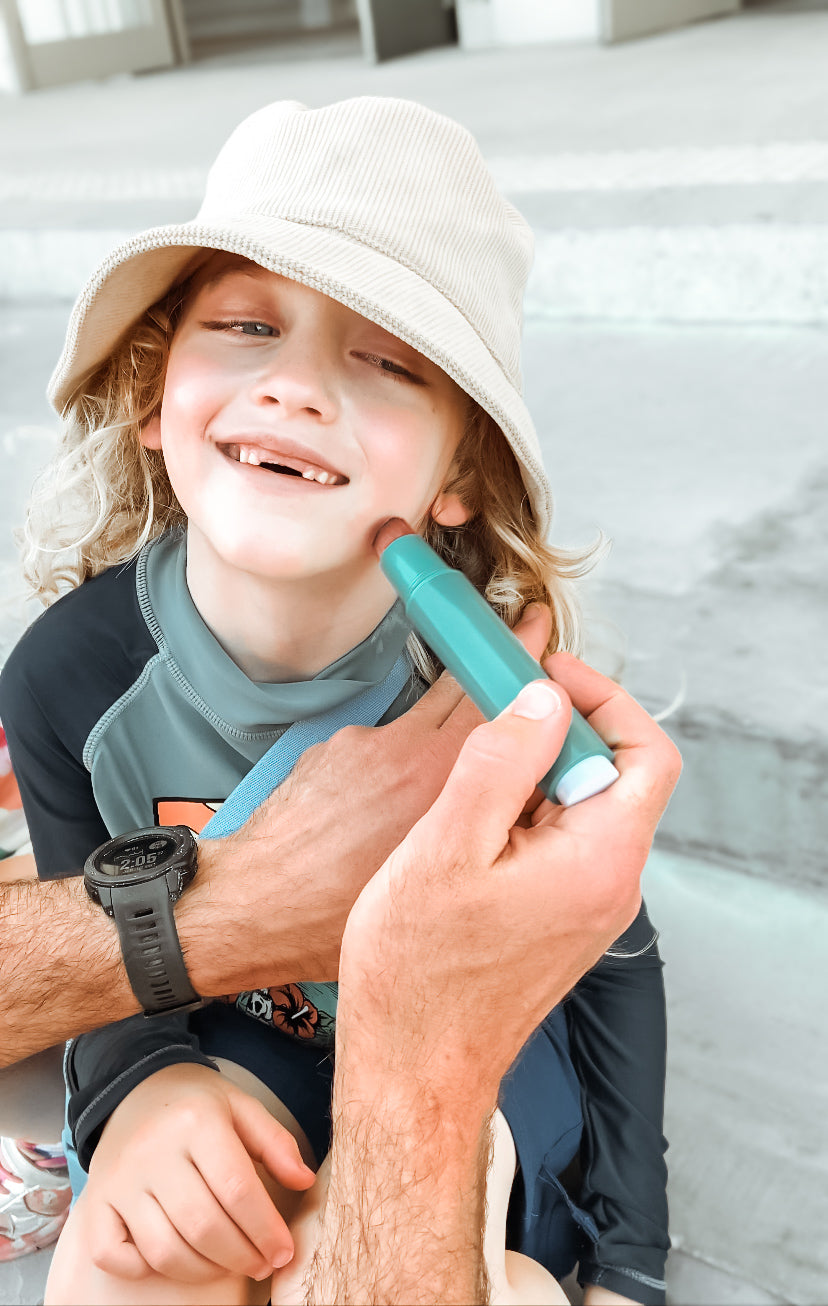 The picture is of a young boy, who appears to be very happy and is smiling widely. The child is wearing a light-colored bucket hat and has long, curly blonde hair. There is an adult hand holding a SUNNYBOD™ Refillable Sunscreen Brush and is applying sunscreen to the child's face. The child is also wearing a dark-colored swim shirt with some design on it. They are on grey steps.