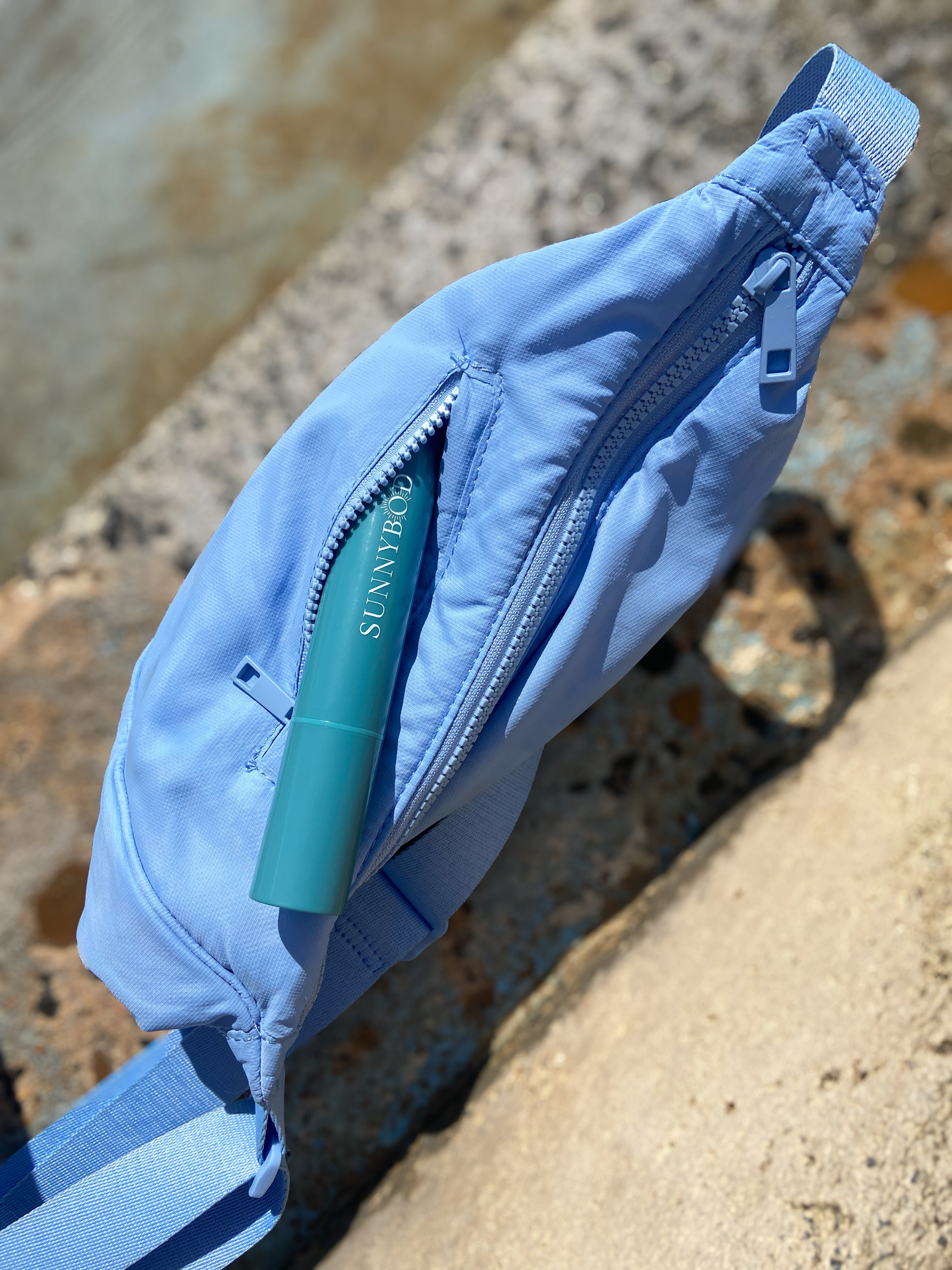 A blue waist bag or fanny pack. The bag is placed on a concrete ledge. The bag is light blue in color with a zipper in the middle. There is a turquoise SUNNYBOD™  Refillable Sunscreen Brush partially sticking out from the zipper pocket. The background of the image is blurred, so the focus is on the bag and sunscreen applicator. 