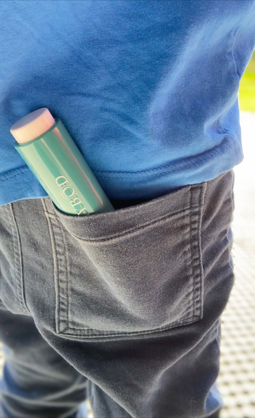 A close up picture of a SUNNYBOD™ Refillable Sunscreen Brush in the pocket of a child's Jean pocket. The jeans are dark blue and the child is wearing a sky blue t-shirt. The susncreen brush is turquoise in colour with a white pump button. The end of the brush is protruding from the jeans pocket.