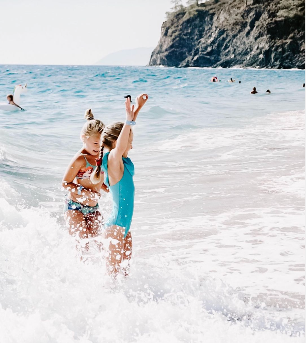 Two girls with their SUNNYBOD refillable sunscreen brush's, playing in the surf at the beach. They are smiling and having fun. 