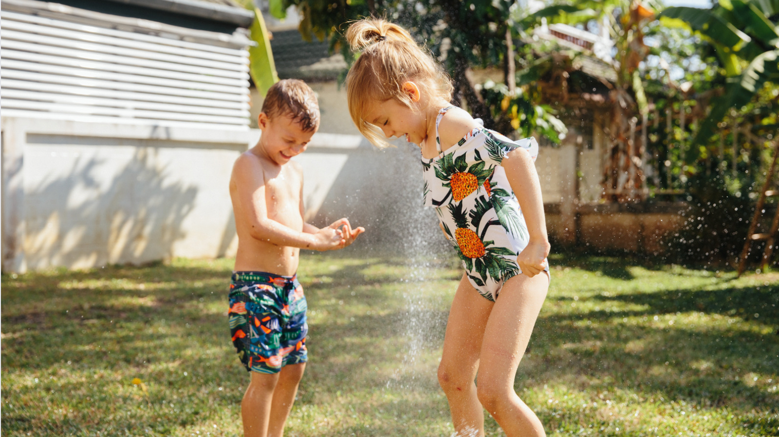 A little boy and girl wearing bright coloured swimwear are playing in a backyard sprinkler on a sunny day. They are smiling and running through the water. 