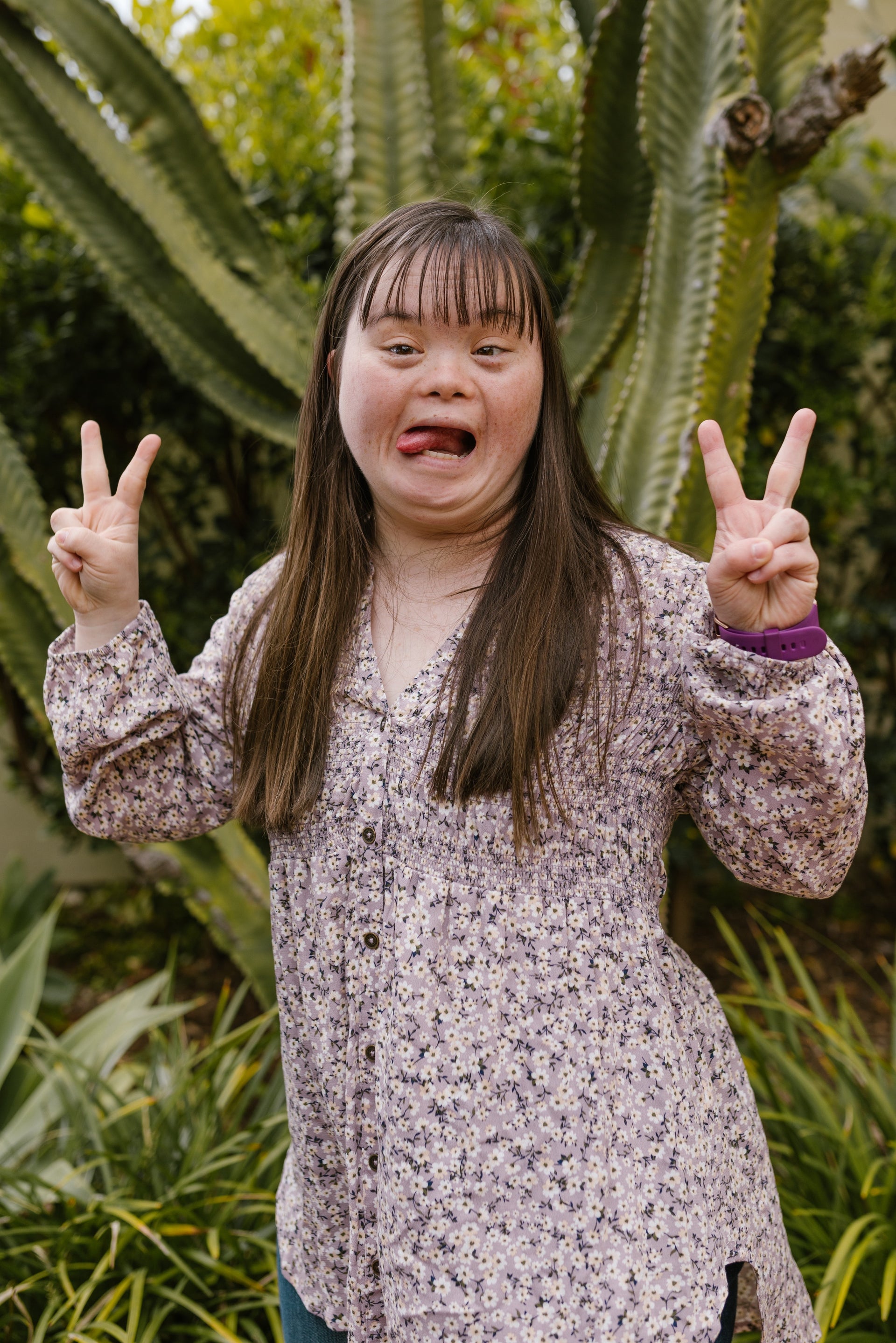 A pretty girl pulls a cheeky face by poking her tongue out. She is holding both hands in the air make the "peace sign" with her fingers. She is standing in a lush garden with a large cactus behind her. 