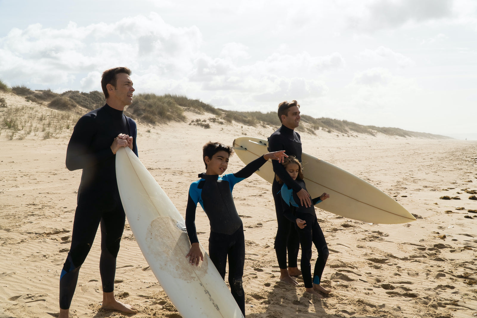 Two dads at the beach with their sons.  They are all wearing wetsuitsThey are holding surfboards and are pointing out to the water. 