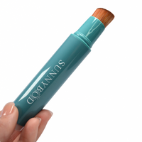 A product picture of a SUNNYBOD™ Refillable Sunscreen Brush Applicator. The brush is turquoise in colour. The susncreen brush bristles are light brown. The handle has the word "SUNNYBOD" written in white capital letters. The brush is placed on a white background and is being help in the fingertips of well manicured hands.